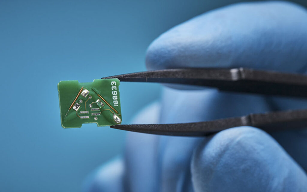 Gloved hand holding a small green circuit board sensor component with tweezers, showing surface-mount components and serial number markings.