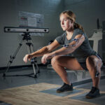 An athlete performing a squat on a platform while an optical tracking system mounted on a tripod captures her movement in a gym environment.