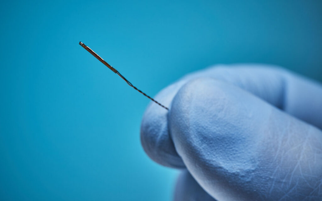 Gloved fingertips holding a miniature electromagnetic sensor coil with a fine twisted lead wire against a blue background.