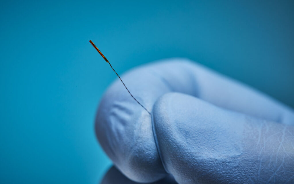 Gloved fingertips holding a miniature electromagnetic sensor coil with a fine twisted lead wire against a blue background.