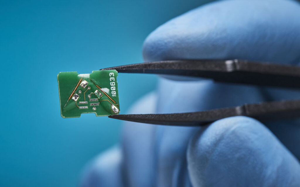 Gloved hand holding a small green circuit board sensor component with tweezers, showing surface-mount components and serial number markings.