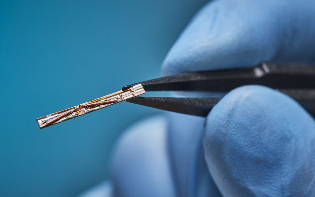 Gloved fingertips holding a miniature electromagnetic sensor coil assembly with exposed copper windings using black tweezers.