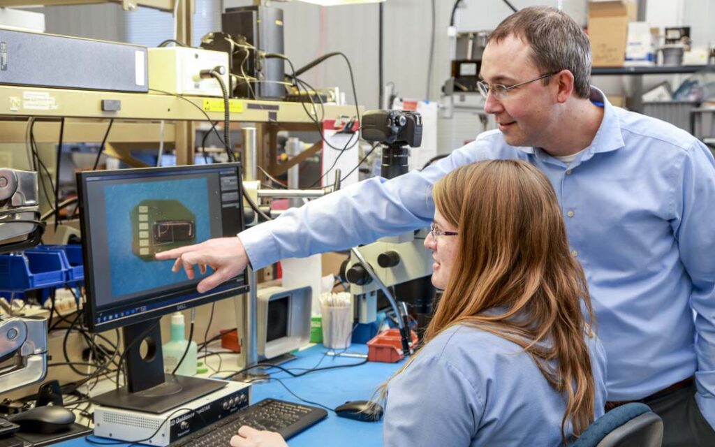 Two NDI employees reviewing a magnified component image on a monitor in a lab, with one pointing at a feature on the screen while the other looks on.