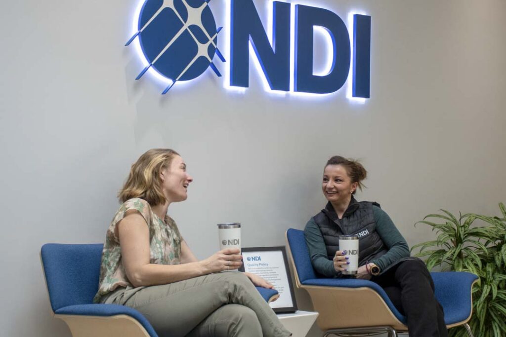 Two NDI employees seated in blue chairs, smiling and chatting while holding NDI-branded tumblers, with the illuminated NDI logo on the wall behind them and a framed Quality Policy displayed on the side table.
