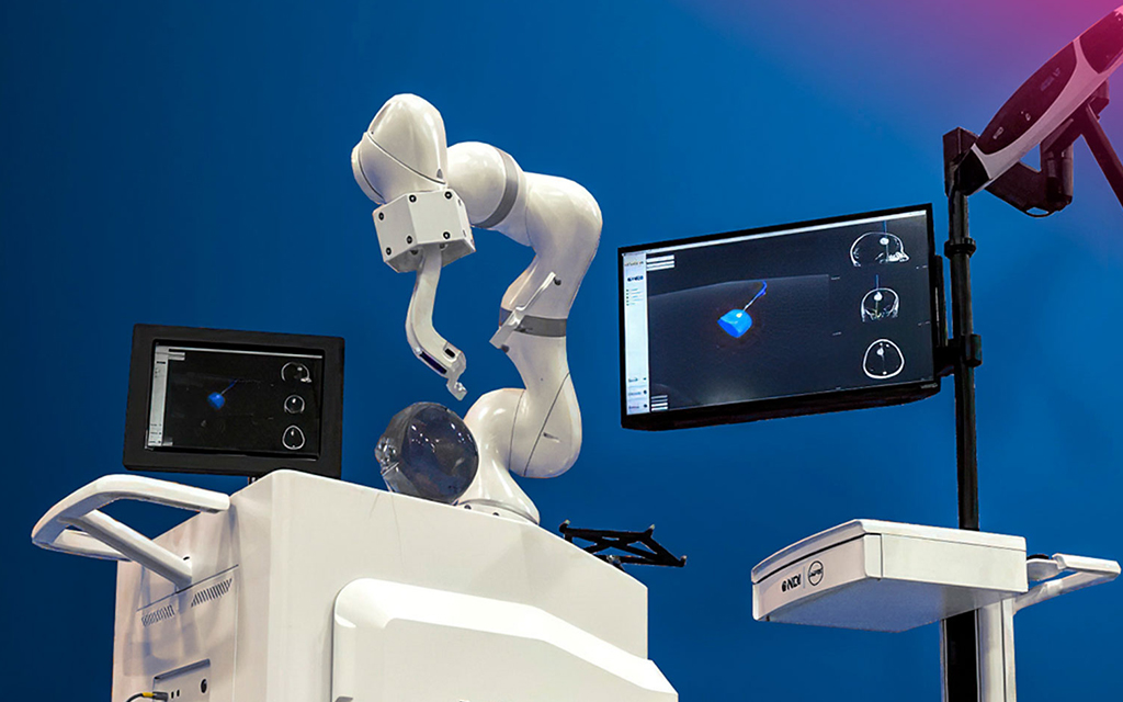 A robotic arm positioned over an anatomical head model, with two monitors displaying tracking visualizations. An NDI Polaris optical tracker is mounted on a stand to the right.