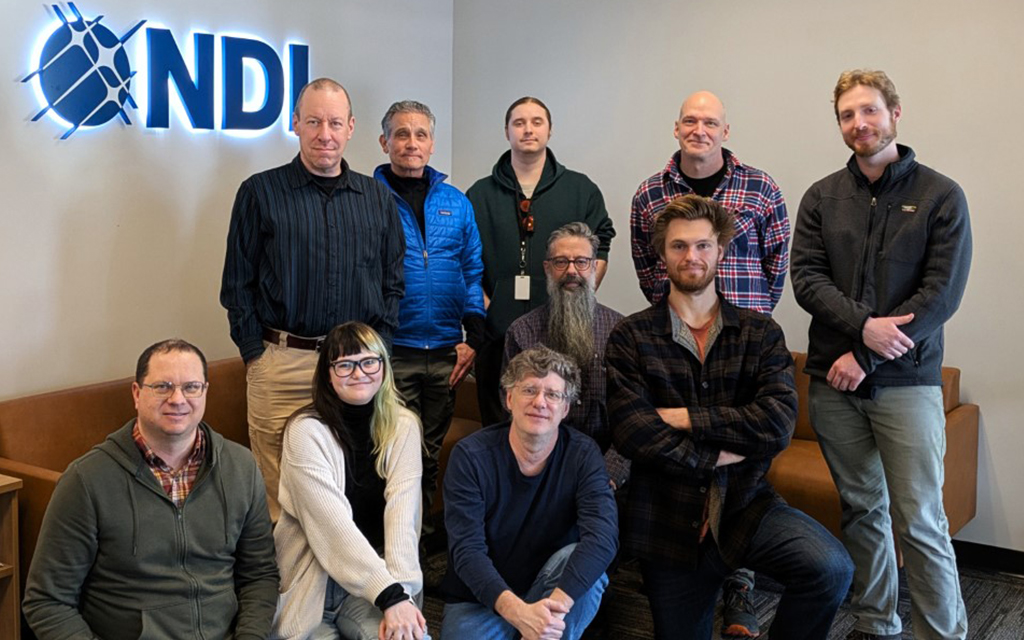 Group photo of an NDI team posing together in front of the illuminated NDI logo, with some team members standing and others seated on a couch.
