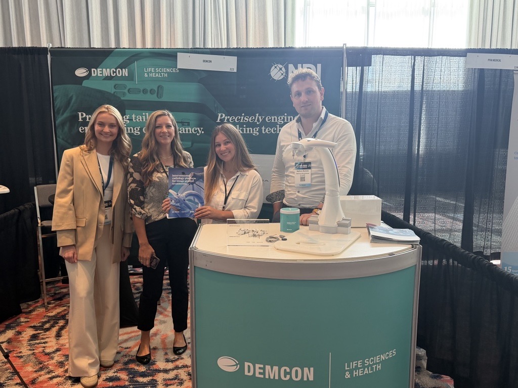 Four representatives at the DEMCON and NDI trade show booth beside a display of tracking technology.
