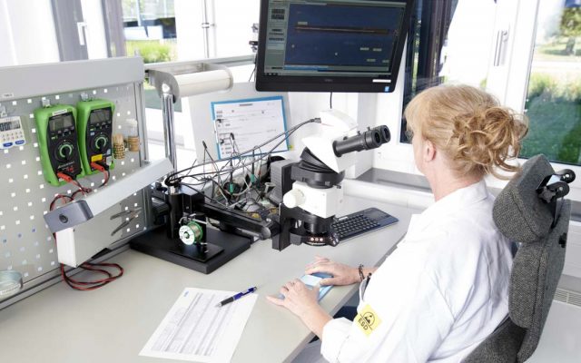 An NDI employee in a lab coat performing quality inspection on a tracking component using a microscope, with multimeters and test equipment at a bright workstation.