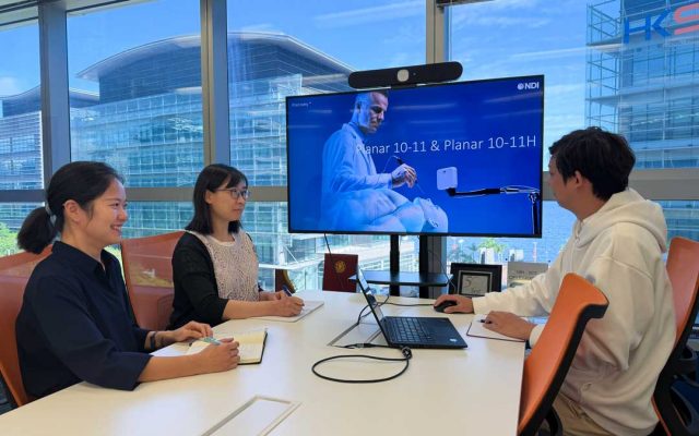 Three colleagues meeting around a conference table, viewing a presentation on a wall-mounted display about NDI's Planar 10-11 and 10-11H tracking systems, with a city skyline visible through floor-to-ceiling windows.