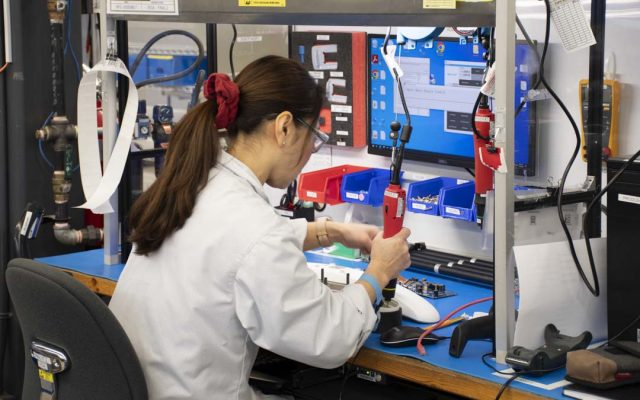 An NDI employee in a lab coat assembling tracking system components at a workstation, with organized parts bins and a monitor displaying assembly instructions.
