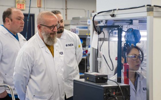 Three NDI employees in lab coats observing a colleague working inside an enclosed manufacturing workstation through a transparent enclosure.